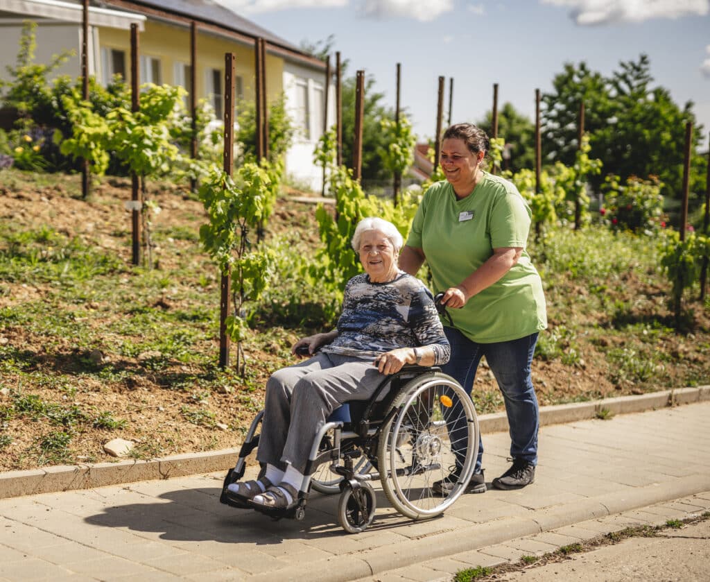 Senioren sitzt im Rollstuhl und wird von Pflegefachkraft durch den Garten geschoben